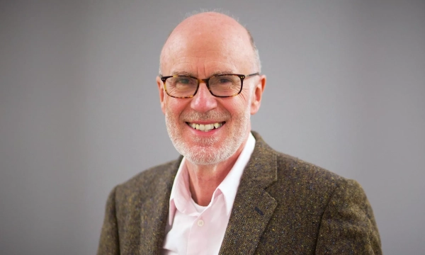 A smiling man with glasses and a bald head, wearing a tweed jacket over a light pink shirt, poses against a neutral background.