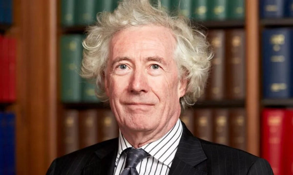 A man with curly gray hair wearing a black suit and striped tie stands in front of shelves filled with colorful books.