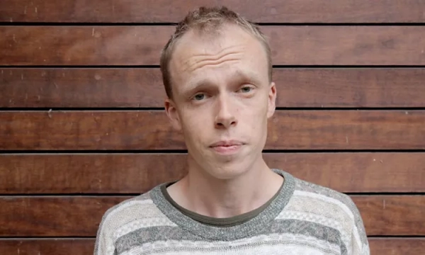 A young man with short hair and a neutral expression stands against a wooden wall, wearing a striped sweater.