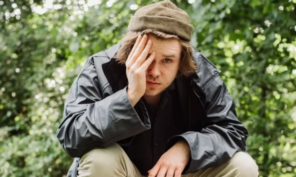 A young man in a green hat and dark jacket sits outdoors, resting his chin on his hand, with a concerned expression against a background of greenery.