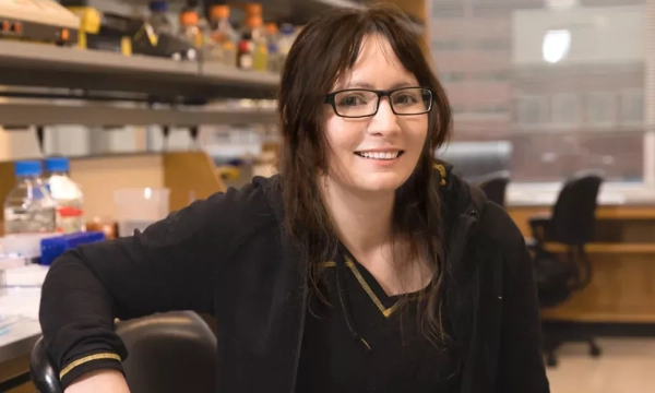 A smiling scientist in a lab, seated with laboratory equipment and containers in the background, wearing glasses and a black hoodie.