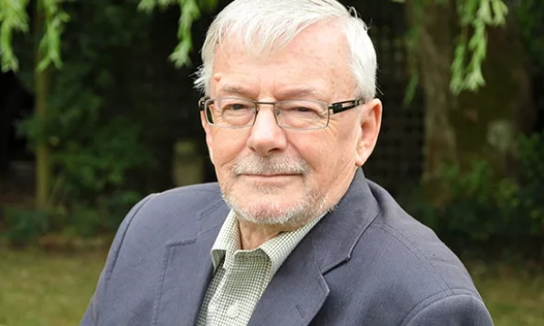 An older man with glasses and gray hair, wearing a blazer, poses outdoors with greenery in the background.