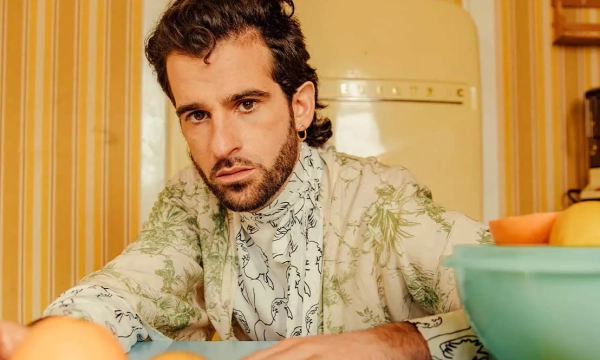 A man with curly hair and a patterned shirt leans over a table, surrounded by bowls of fruit, against a vintage kitchen backdrop.