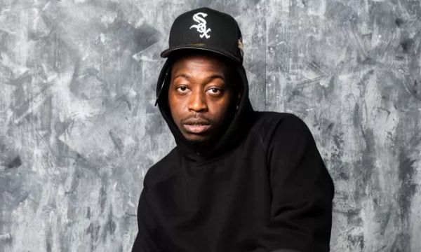 A man wearing a black hoodie and a black baseball cap poses against a gray textured backdrop.