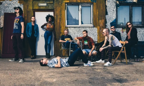 A diverse group of people poses in an urban setting, featuring a mix of fashionable clothing styles, with some seated and others standing against a colorful, textured background.