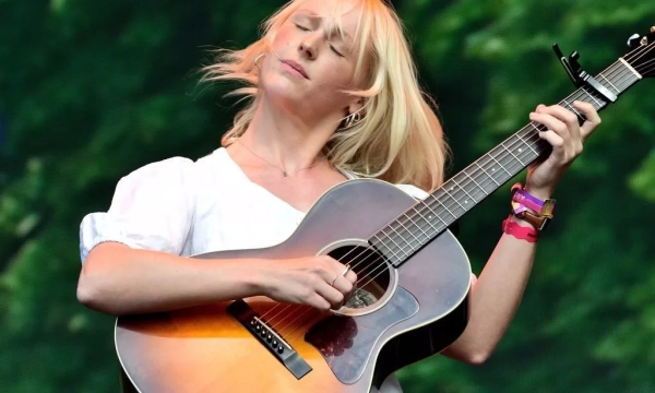 A blonde female musician passionately playing an acoustic guitar on stage, with flowing hair and a vibrant natural background.