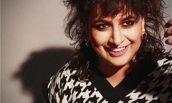 Smiling person with curly hair, wearing a patterned black and white sweater, looking towards the camera against a neutral background.