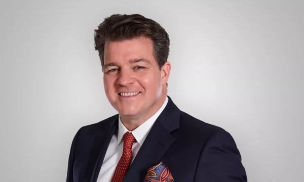 A smiling man in a dark suit and red tie, with a colorful pocket square, poses against a light background.