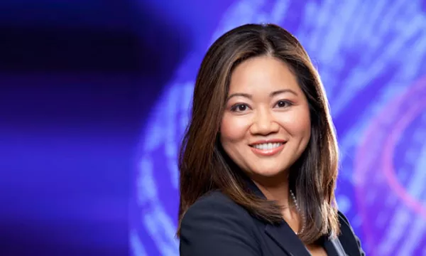 A professional woman with long brown hair smiles confidently against a purple and blue abstract background. She wears a dark blazer.
