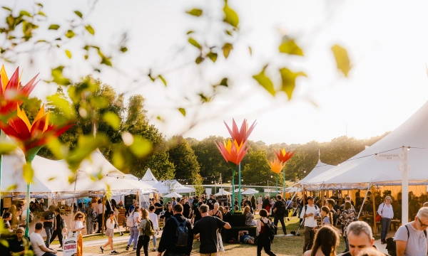 A vibrant outdoor festival scene with large flower sculptures, crowds of people mingling, food stalls, and white tents under a bright sky.
