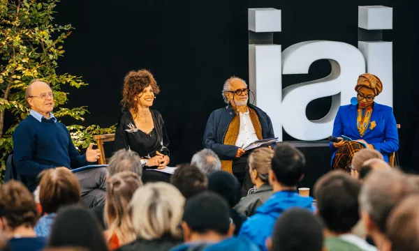 A panel discussion featuring four speakers, with a diverse audience in front, set against a backdrop displaying the letters 