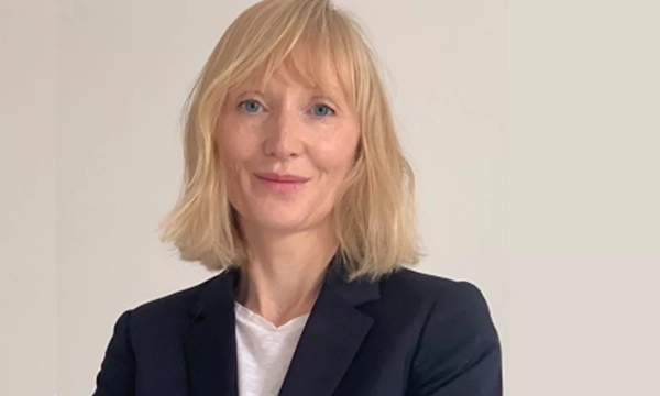 A professional individual with short blonde hair wearing a black blazer and white shirt, standing confidently against a plain background.