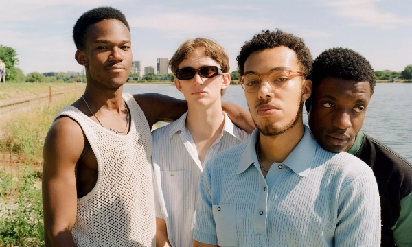A group of four young men stands by a river, dressed casually in summer attire, with sunglasses and a bright sky in the background.
