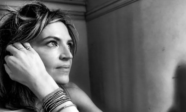 A contemplative woman with styled hair resting her hand on her face, wearing multiple bracelets, set against a softly lit background.