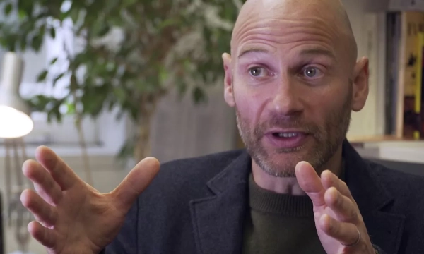 A man with a bald head and beard gestures passionately while speaking in an office setting, surrounded by plants and books.