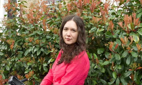 A young woman with long, wavy hair sits outdoors against a backdrop of green and reddish leaves, wearing a bright pink sweater.