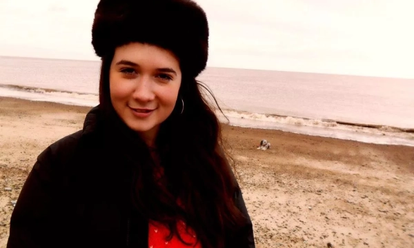 A woman with long hair wearing a fur hat and a black jacket stands on a beach, smiling at the camera, with a calm sea in the background.
