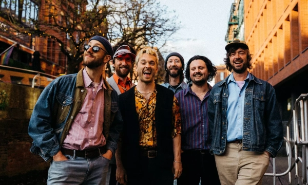 A group of six men dressed in vibrant and eclectic clothing, smiling and posing together in a lively urban setting, with trees and buildings in the background.