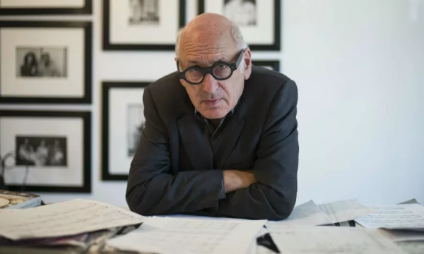 A bespectacled man in a dark suit leans on a cluttered desk covered with papers, set against a backdrop of framed black-and-white photographs.