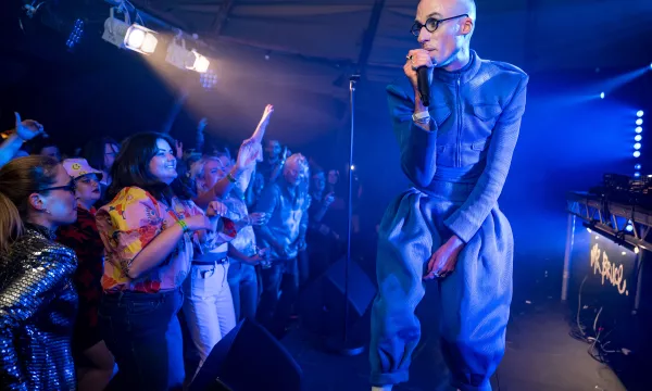 A performer in a blue outfit sings into a microphone on stage, engaging a lively audience of fans in colorful clothing.