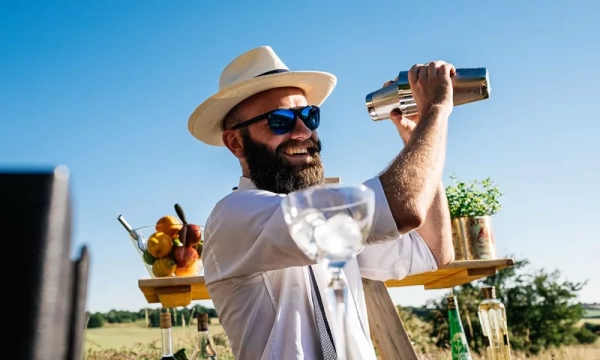 A smiling bartender with a beard and sunglasses shakes a cocktail shaker, wearing a white shirt and straw hat, with a bar setup in the background.