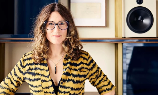 A woman with curly hair and glasses stands confidently in front of shelves with speakers and framed documents, wearing a yellow and black patterned shirt.