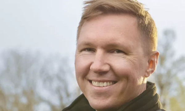 A smiling man with short, light brown hair wearing a black jacket, standing outdoors with trees blurred in the background.