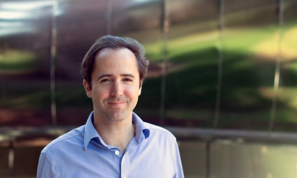 A smiling man wearing a blue shirt stands in front of a reflective metallic surface.