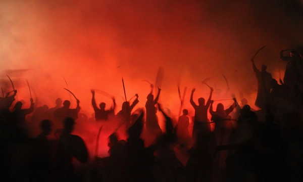 A chaotic scene of silhouetted figures in a fiery atmosphere, wielding weapons and raising their arms amid smoke and orange lighting, suggesting a protest or conflict.