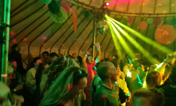 A vibrant crowd dancing in a lively tent, illuminated by colorful lights and a disco ball, creating a festive atmosphere.