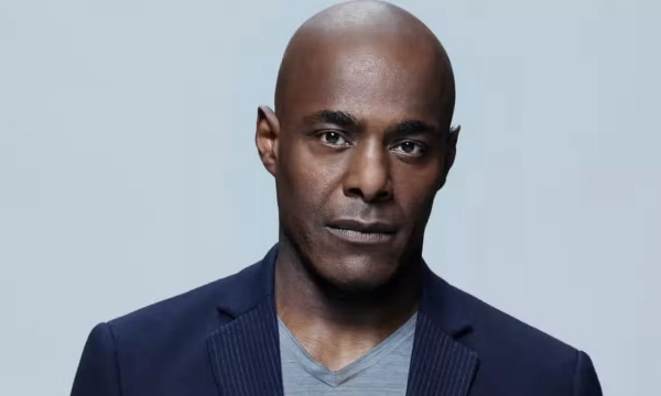 A man with a bald head and a serious expression, wearing a dark jacket over a gray shirt, against a neutral background.