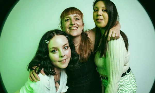 Three women smiling and posing together in front of a soft green background, showcasing a cheerful and friendly atmosphere.