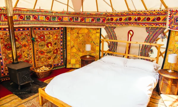 A cozy, brightly decorated yurt interior featuring a wooden bed with white bedding, colorful wall tapestries, a stove, and bedside tables.