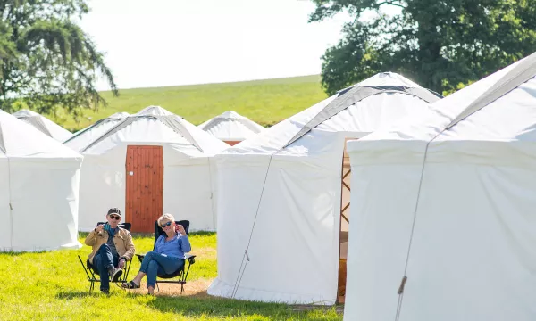 Two people relax in folding chairs outside white yurt-style tents on a grassy field, with green hills in the background.