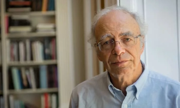 A thoughtful older man in a light blue shirt stands in front of a bookshelf filled with books in a well-lit room.