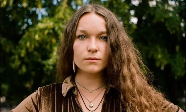A woman with long, wavy hair stands confidently outdoors, wearing a velvet shirt and layered necklaces, with trees and soft natural lighting in the background.