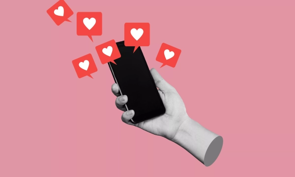 A hand holding a smartphone with heart reaction icons floating above it, set against a pink background, symbolizing social media engagement and appreciation.