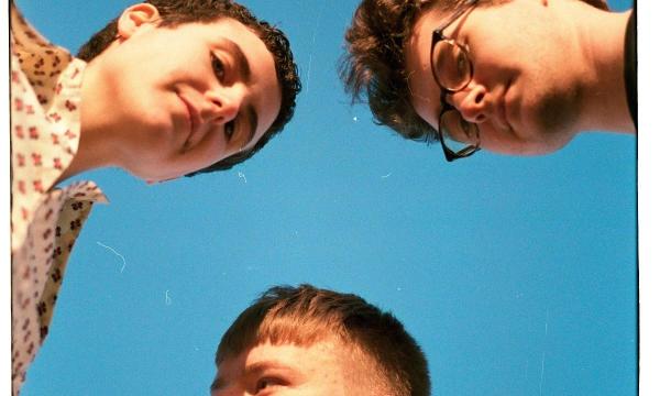 Three young people are seen from below, looking down at the camera against a bright blue sky, showcasing varied hairstyles and clothing styles.