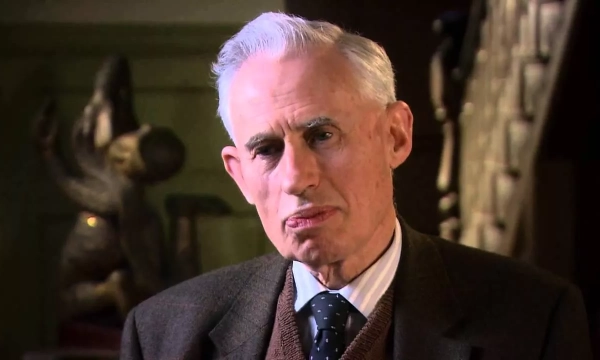 Elderly man in a tweed suit with grey hair, sitting against a blurred background displaying sculptures and art, appearing contemplative.