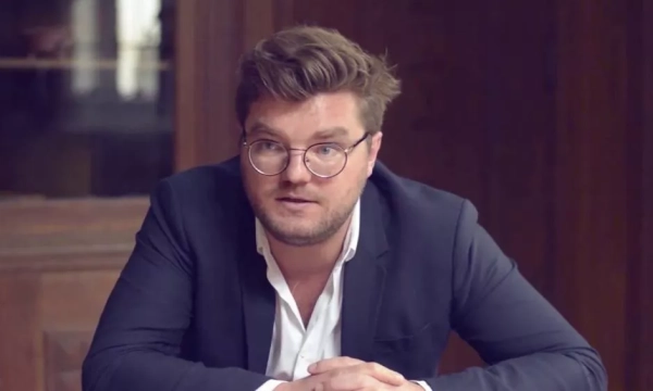 A man with glasses and a stylish haircut sits at a table, wearing a dark suit and white shirt, with a serious expression in an elegant room.