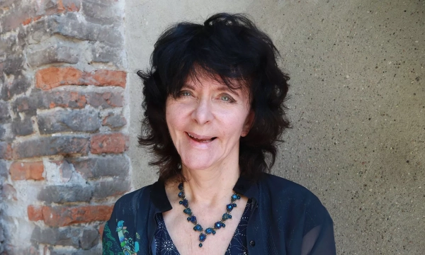 A woman with dark, wavy hair smiles warmly against a textured stone wall, wearing a decorative necklace and a patterned top.