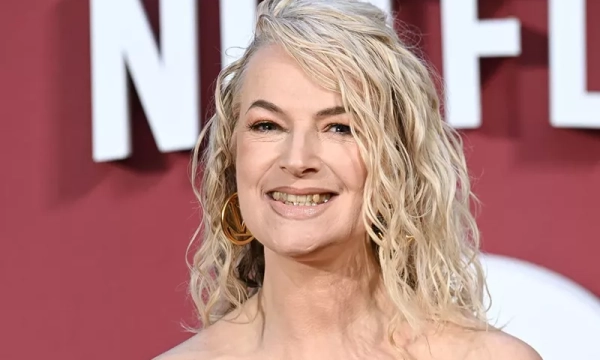 A smiling woman with curly blonde hair and large hoop earrings poses in front of a red Netflix backdrop at a public event.