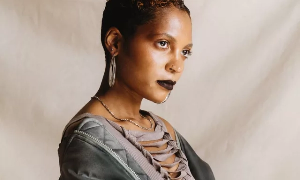 A person with short, styled hair and dark lipstick poses against a neutral background, wearing a trendy outfit with a unique design.
