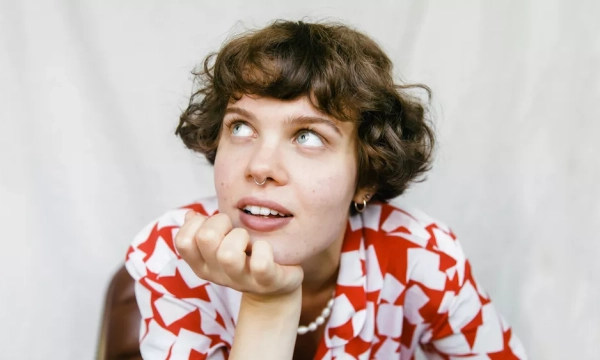 A young person with short, curly hair and a thoughtful expression rests their chin on their hand, wearing a patterned red and white outfit against a light background.
