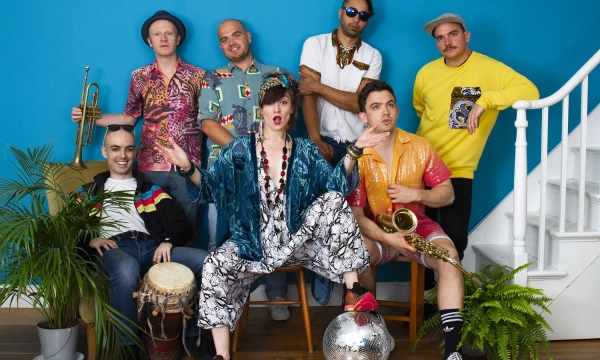 A lively group of seven musicians poses together in colorful outfits, with a mix of instruments including a trumpet and saxophone, set against a bright blue wall.