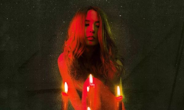 A woman with wavy hair sits amidst glowing candles, surrounded by a dark, starry background that creates an ethereal atmosphere.