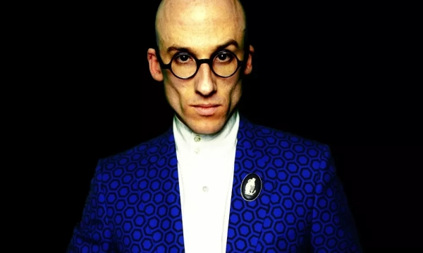 A bald man in a patterned blue suit and round glasses poses against a dark background, exuding confidence with a serious expression.