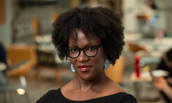 A woman with natural curly hair and glasses, wearing earrings, posing confidently in a bright, modern indoor setting.