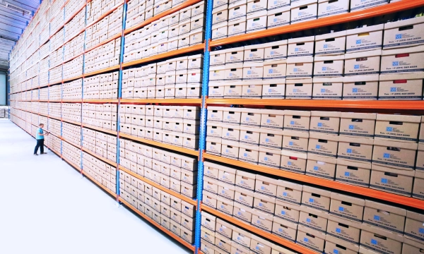 A large warehouse filled with neatly stacked boxes on towering shelves, with a person standing beside them for scale.