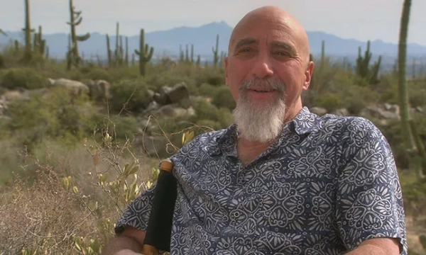 A man with a beard and a patterned shirt sits in a desert landscape, featuring cacti and mountains in the background.